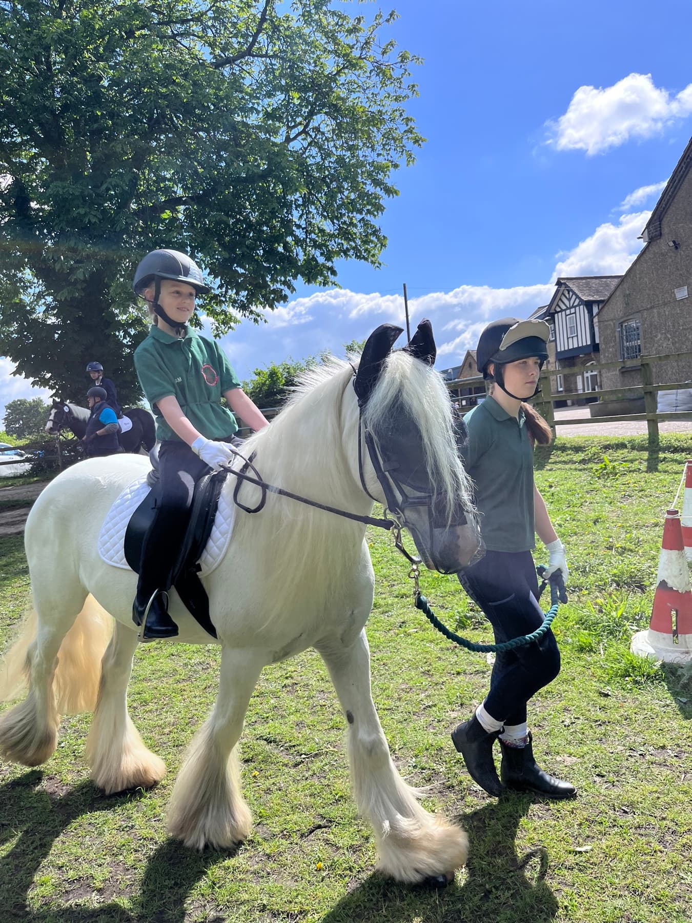 A young RDA rider on a grey cob in the sunshine, with a volunteer walking beside them holding the lead rein.