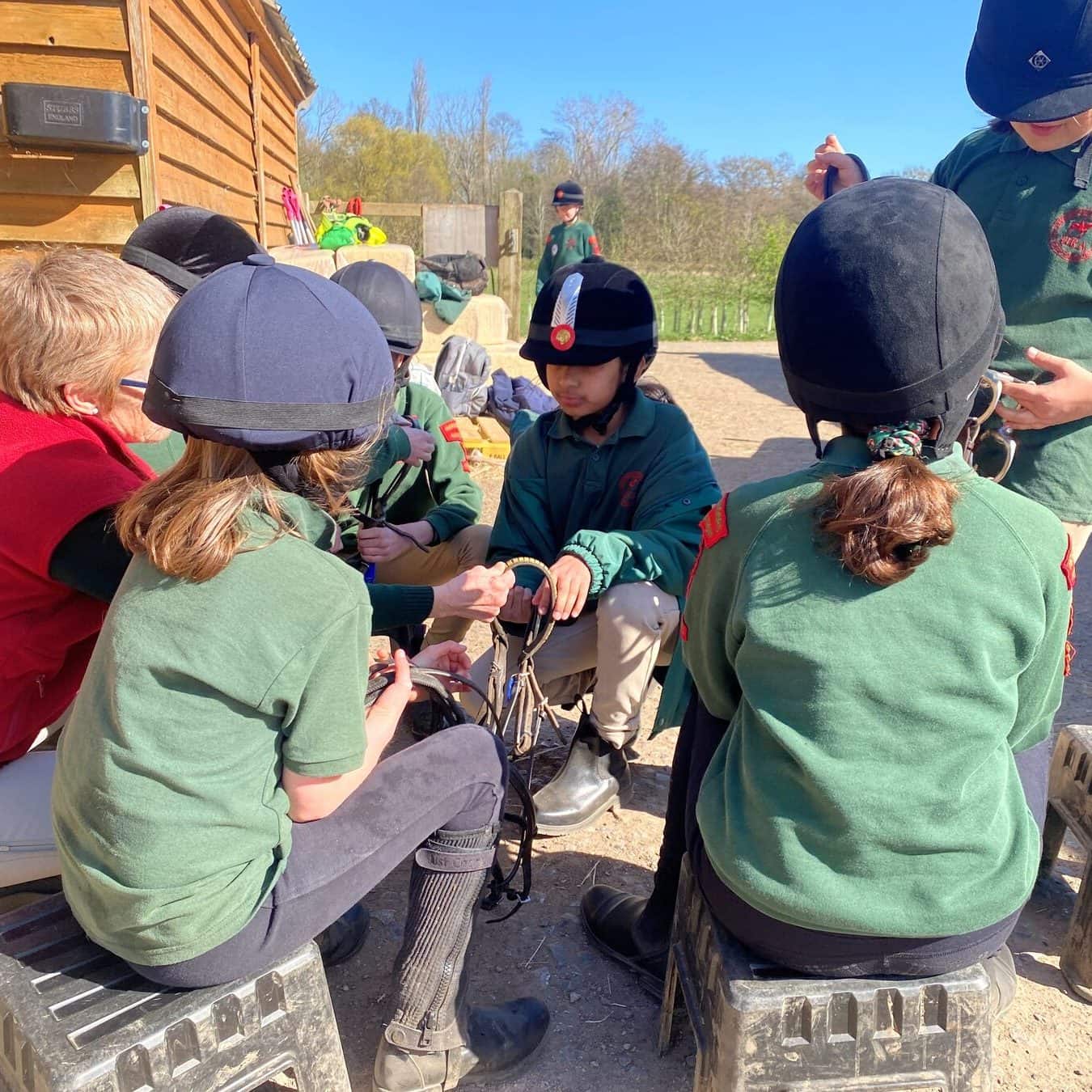 An adult sitting with a group of young Horse Rangers beside the hay barn in the sunshine, while they work on a bridle together