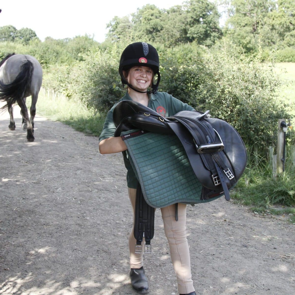 A Horse Ranger grinning as they carry a saddle and numnah along the track, with a pony being led ahead