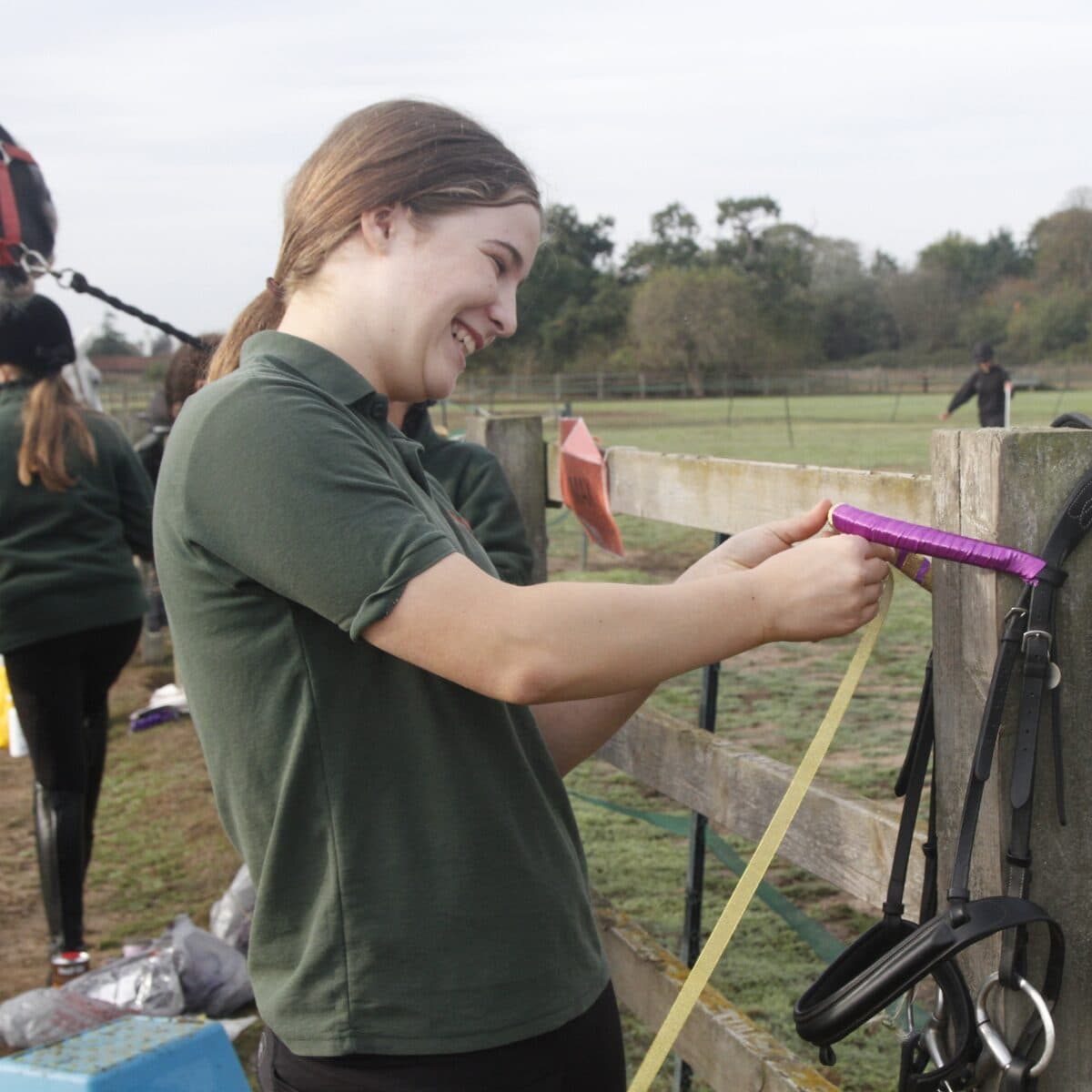 A Horse Ranger laughing as she winds purple tape around a bridle hung on the paddock fence, with other Rangers working behind her