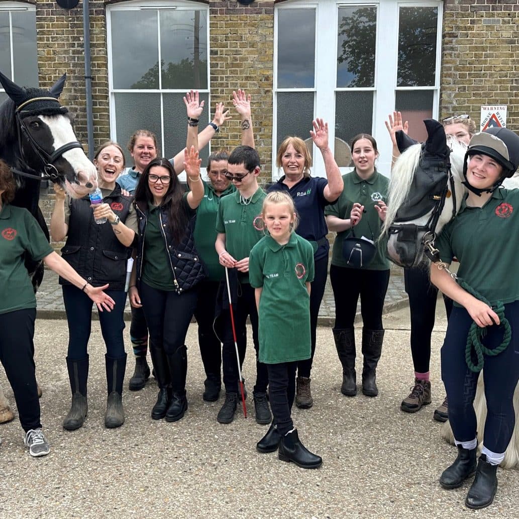 RDA riders, volunteers and coaches waving together in the yard, two riders mounted on a black and white cob and a grey.